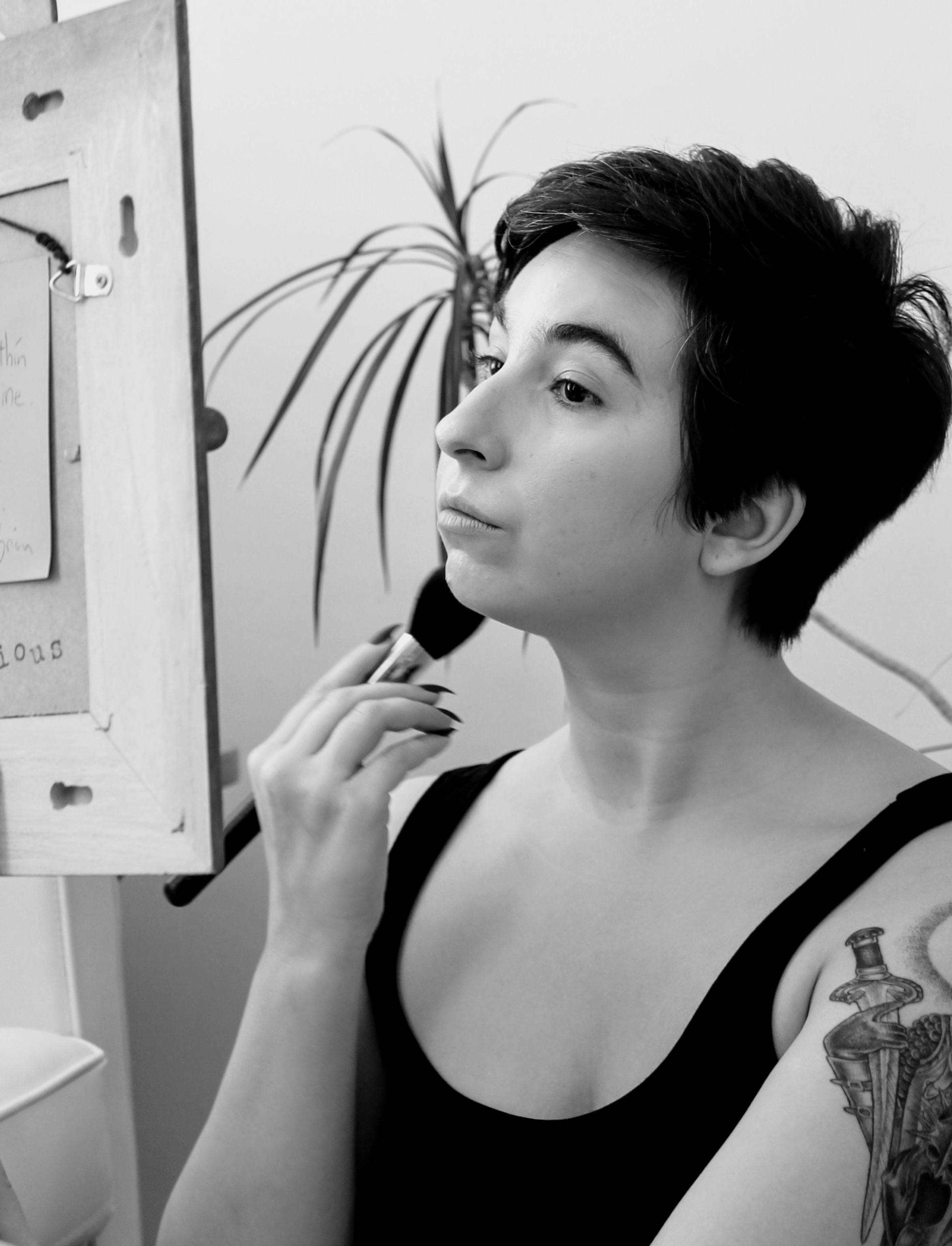 A black and white photo of
                        Cara Marta Messina. She is a white, cis woman with short brown hair. She is looking in a mirror putting on blush. She has a tattoo on her left shoulder of Eowyn from Lord of The Rings.